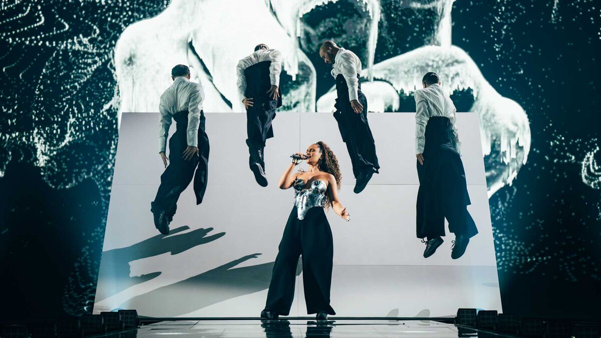 Alicja Szemplińska występuje na scenie z mikrofonem, a wokół niej znajdują się tancerze w białych koszulach i czarnych spodniach. Za nimi widać dużą wizualizację oraz biały element scenografii.