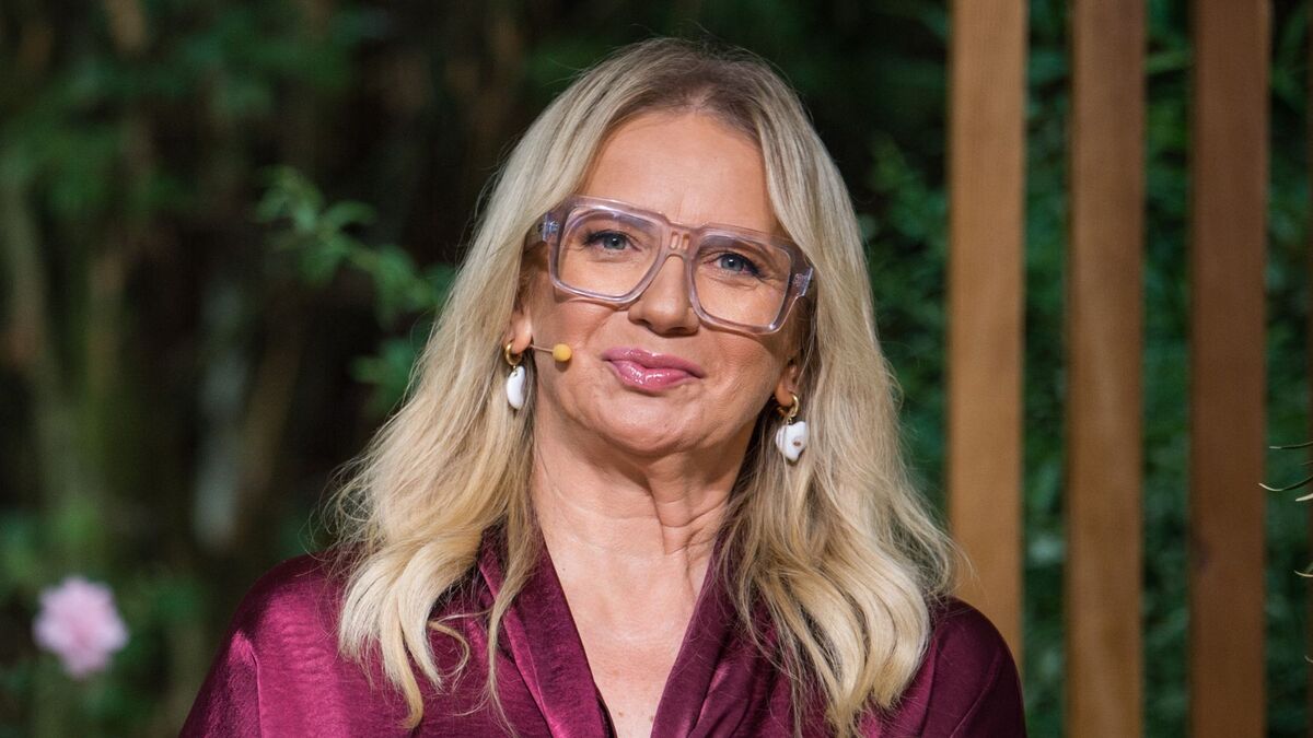 Marzena Rogalska ma długie blond włosy, duże transparentne okulary i mikrofon przy policzku. Ubrana jest w satynową bluzkę w kolorze bordowym oraz białe, wiszące kolczyki. W tle widać rozmytą zieleń roślin i pionowe drewniane elementy scenografii.