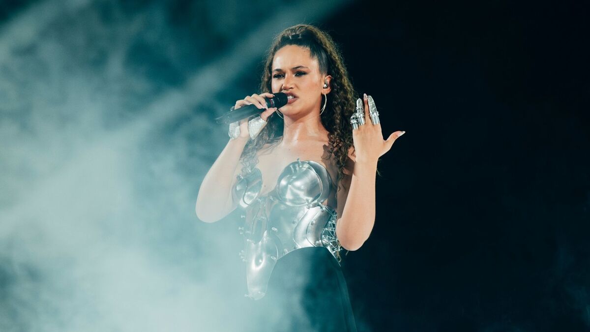 Alicja Szemplińska śpiewa na scenie, trzymając mikrofon w dłoni. Wokalistka ma srebrną, metaliczną stylizację oraz wysokie, kręcone włosy związane w kucyk. Wokół artystki widoczna jest sceniczna mgła i ciemne tło oświetlone reflektorami.