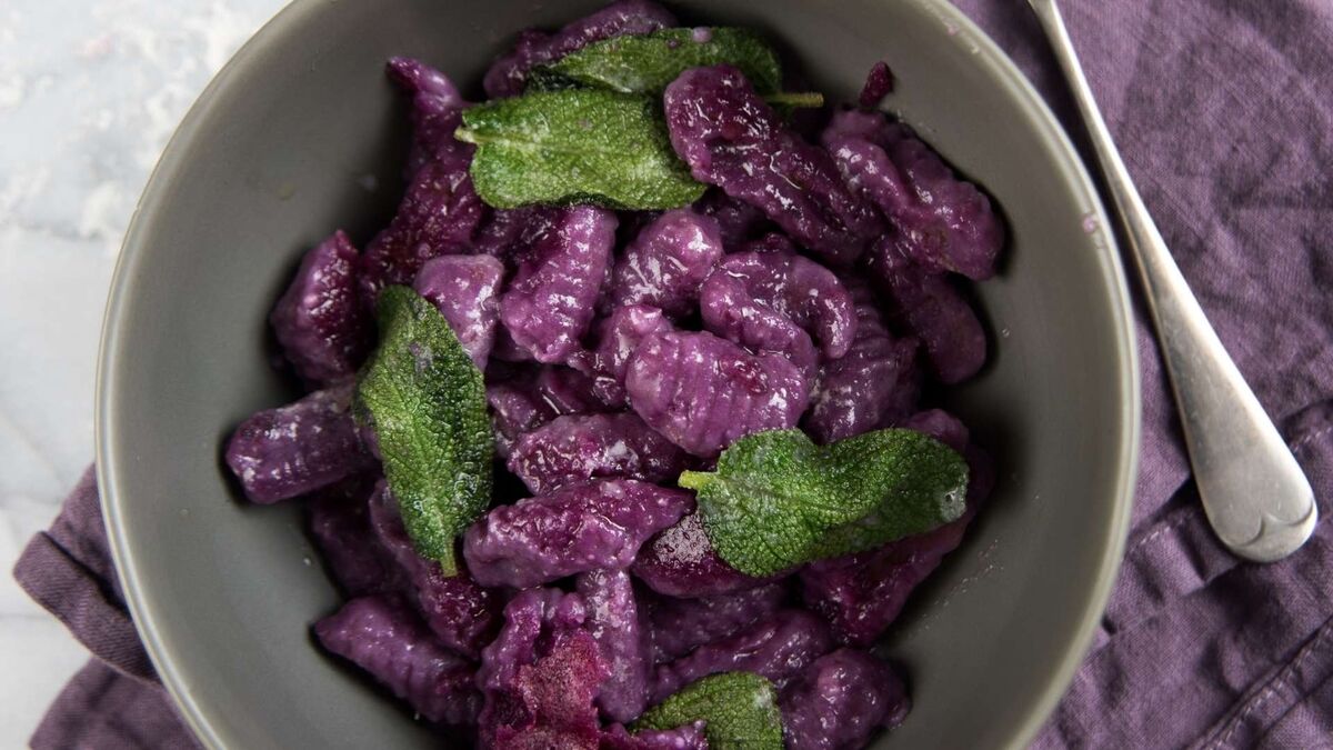 Miska wypełniona fioletowymi gnocchi, podanymi z listkami świeżej szałwii. Kluseczki mają intensywny, purpurowy kolor i delikatnie połyskującą powierzchnię. Obok naczynia leży łyżka, a całość ustawiona jest na jasnym blacie z fioletową serwetką.