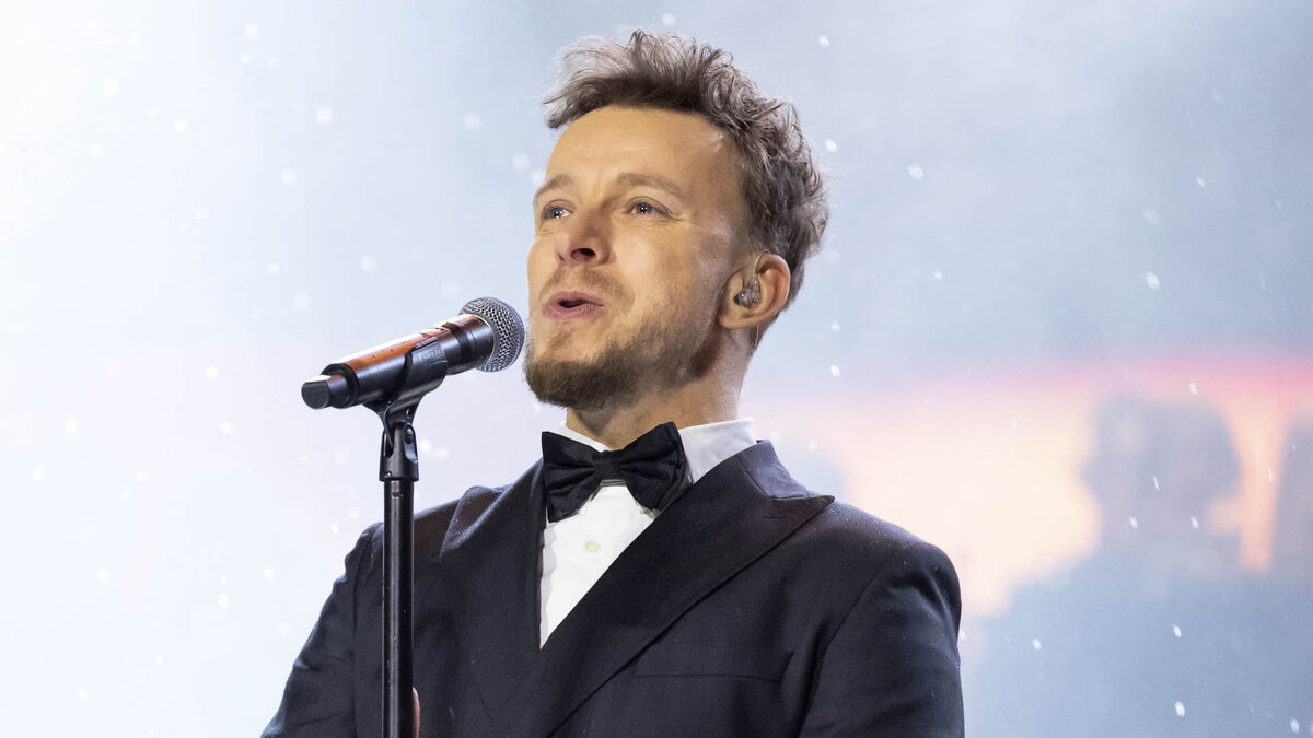 Piotr Kupicha w czarnym smokingu i muszce śpiewa do mikrofonu na statywie podczas występu scenicznego. Na jego uchu widać odsłuch douszny. W kadrze widoczne są drobne, białe płatki lub konfetti opadające na tle jasnej scenografii.