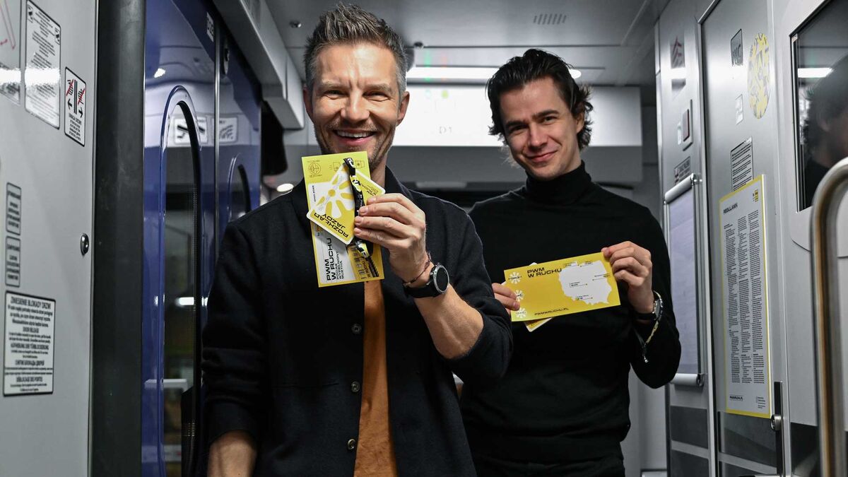 Pociąg wypełniony muzyką. Na zdjęciu w korytarzu pociągu stoi Adam Sztaba i drugi mężczyzna , którzy prezentują żółte materiały promocyjne. W tle widać drzwi i oznaczenia na ścianach wagonu oraz elementy wyposażenia przedziału.