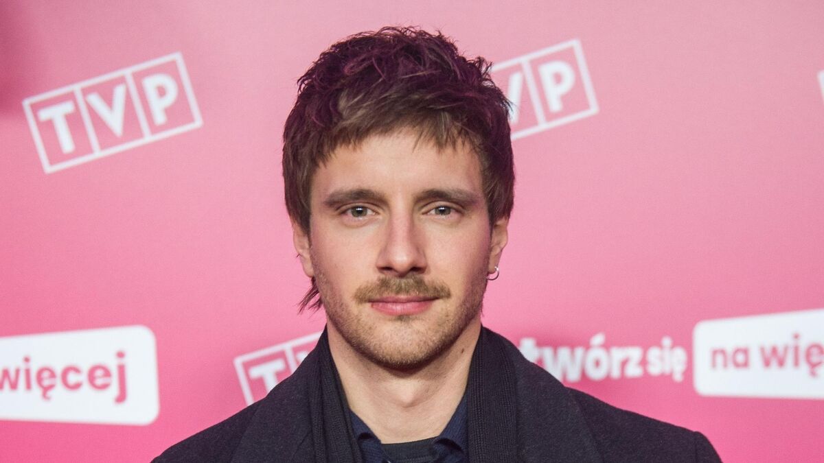 Maciej Musiał pozuje do zdjęcia na tle ścianki z logo TVP. Ma na sobie ciemny płaszcz i ciemną koszulę oraz niewielki kolczyk w lewym uchu. Fotografia została wykonana podczas wydarzenia promocyjnego przed różowym tłem z logotypami.