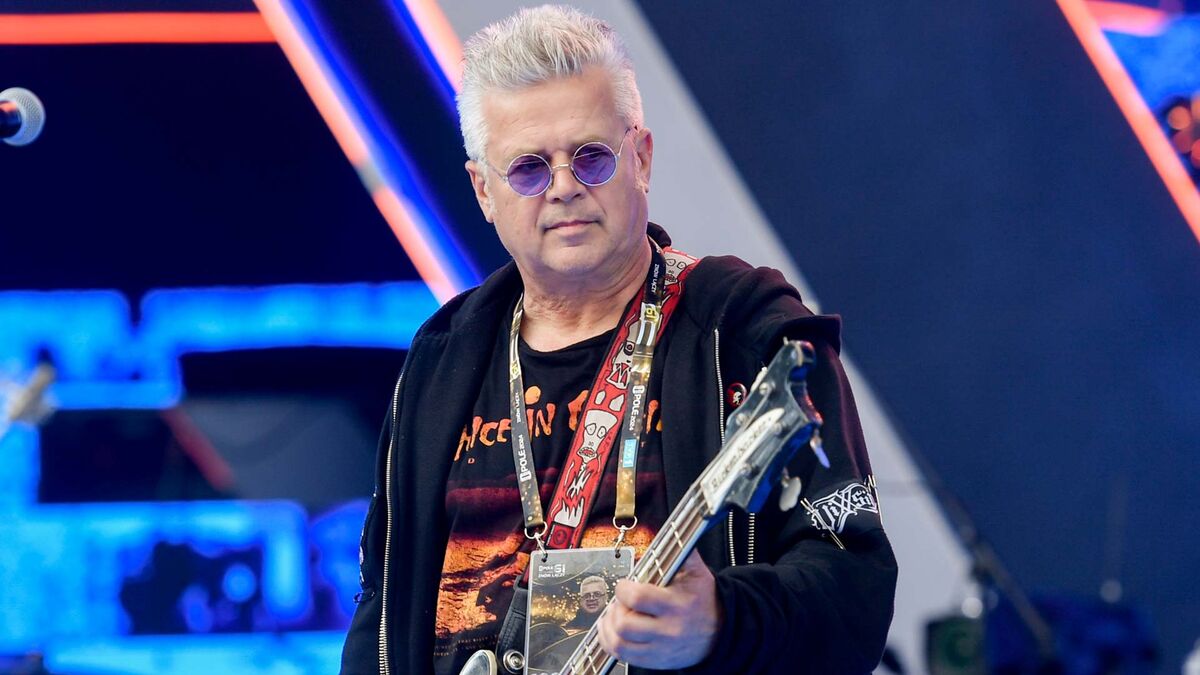 Jacek Jędrzejak gra na gitarze basowej, trzymając instrument przed sobą. Ma siwe włosy, okrągłe okulary z fioletowymi szkłami, czarną bluzę i koszulkę z nadrukiem. W tle znajdują się elementy sceny oraz niebieskie i czerwone światła.