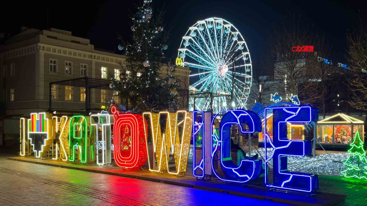 Na placu w Katowicach widać podświetlany napis „#KATOWICE” w kolorowych neonach. W tle stoją choinka z lampkami oraz diabelski młyn. Ujęcie wykonano nocą, a iluminacje odbijają się na nawierzchni.