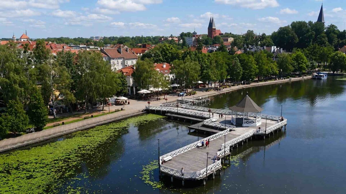 Panoramiczny widok przedstawia miasto położone nad jeziorem z drewnianym pomostem prowadzącym do zadaszonej altany. Wzdłuż nabrzeża widoczne są drzewa, zabudowa z czerwonymi dachami oraz spacerujący ludzie, a na wodzie unoszą się liście lilii wodnych. W tle znajdują się wieże kościołów oraz rozległa panorama miasta pod błękitnym niebem z chmurami.