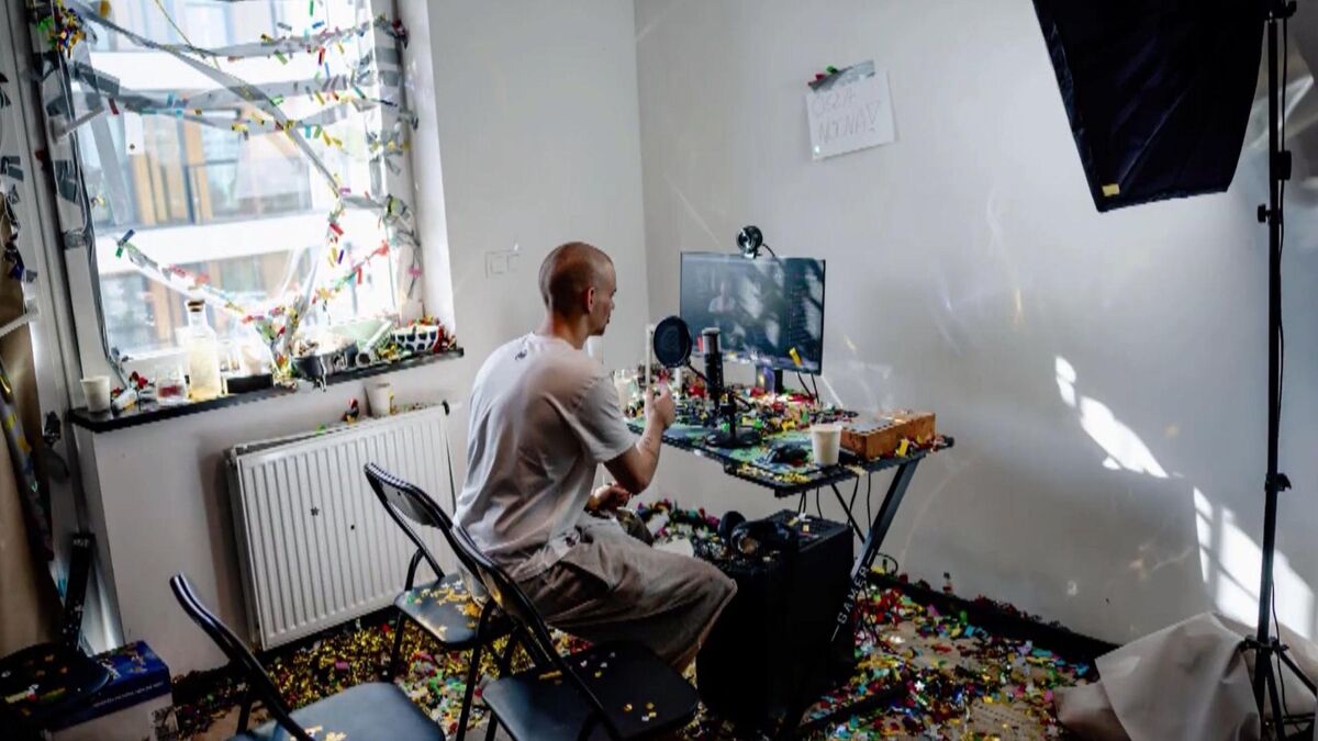 W pomieszczeniu stoi biurko z komputerem, mikrofonem i kamerą internetową ustawioną nad monitorem. Na podłodze, parapecie i wokół stanowiska rozsypane są kolorowe klocki oraz konfetti. Przy biurku siedzi Łatwogang w jasnym ubraniu, a obok ustawiono lampę studyjną i składane krzesła.